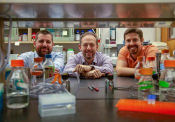 Researchers Antoine Louveau, Jonathan Kipnis and Sandro Da Mesquita smile while peering through lab shelves. There are colorful bottles and other equipment in the foreground.