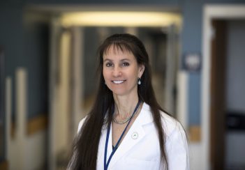 UVA neurologist Karen Johnston, MD, smiles while wearing a white lab coat in front of a blurry hallway.