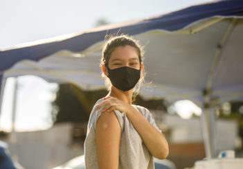 A teen girl wearing a mask shows her small, round vaccination bandage.