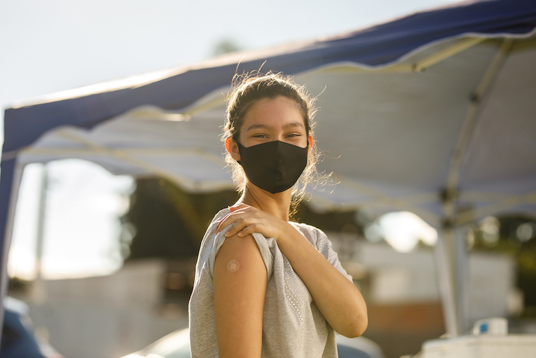A teen girl wearing a mask shows her small, round vaccination bandage.