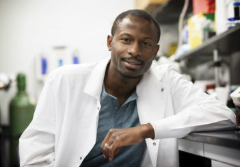 Ukpong B. Eyo leans on a lab bench while wearing a white coat.