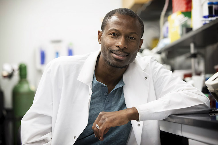 Ukpong B. Eyo leans on a lab bench while wearing a white coat.